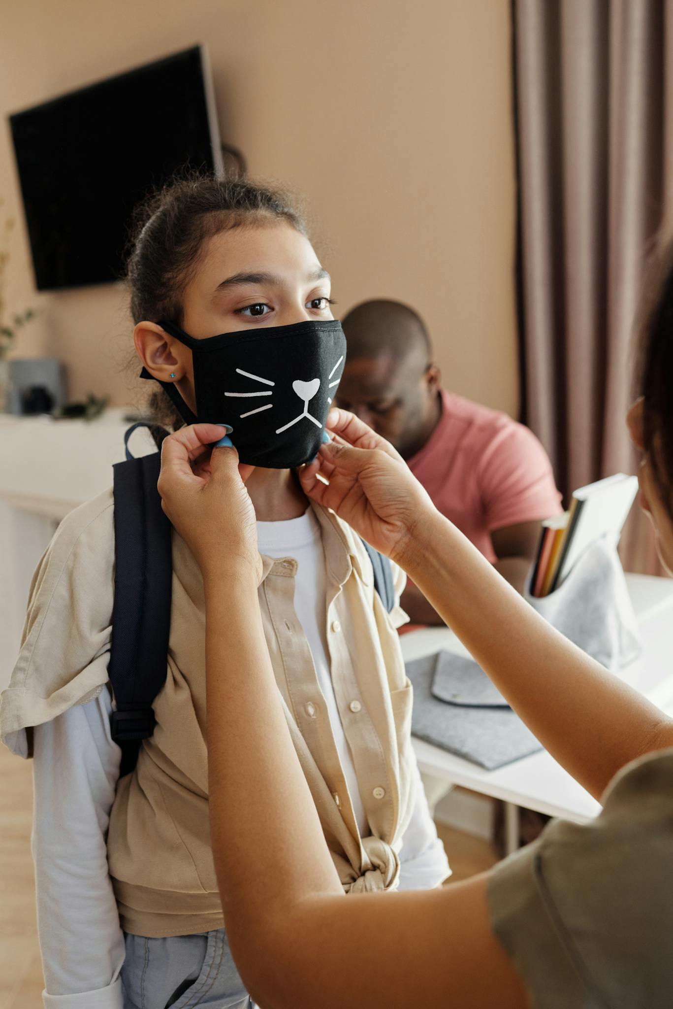 A mother helps her daughter with a cat-pattern face mask indoors, ensuring safety.