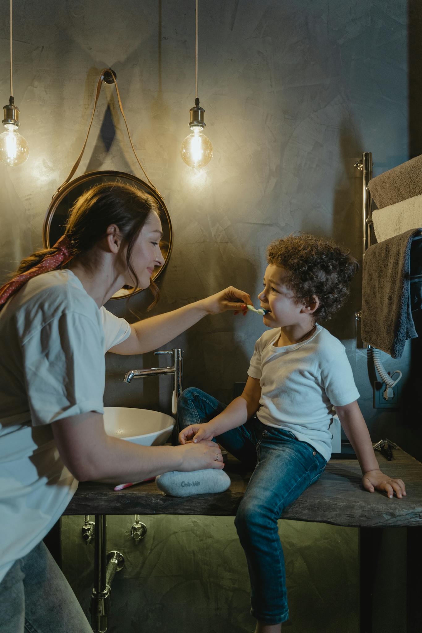 A mother helps her son brush his teeth in a stylish bathroom setting, under warm lighting.